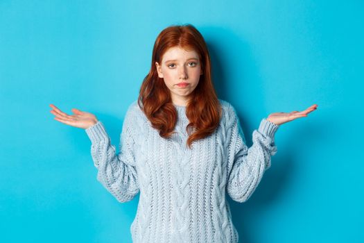 Clueless redhead girl shrugging and saying sorry, standing puzzled against blue background, have no idea.