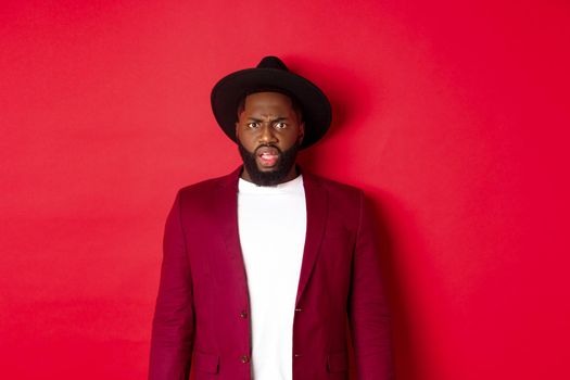 Fashion and party concept. Confused Black man in classy outfit frowning and staring at camera displeased, cant understand, standing over red background.