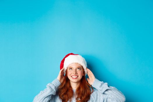 Winter holidays and Christmas eve concept. Head of cute redhead girl in santa hat, appear from bottom and looking up at copy space, staring logo, standing over blue background.