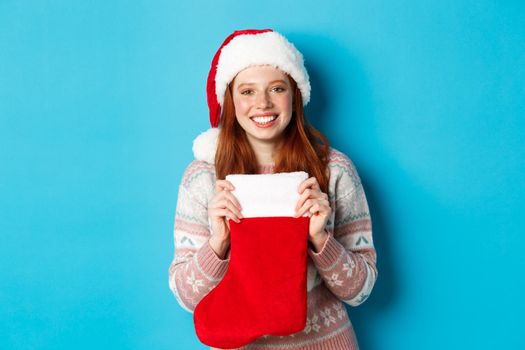 Winter and holidays concept. Cheerful girl receiving presents in Christmas stocking, wearing santa hat and smiling happy, standing over blue background.