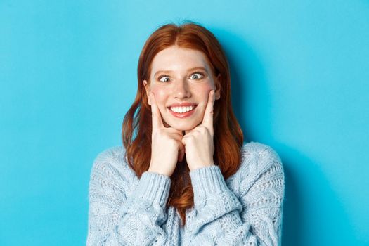 Close-up of funny redhead teen girl making faces, squinting and squeezing cheeks, standing against blue background.