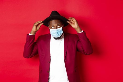 Covid-19, quarantine and holidays concept. Handsome and stylish african american man in face mask, put hat on head and looking sassy, dress-up for party, standing over red background.