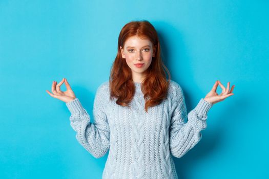 Smiling confident girl with red hair staying patient, holding hands in zen, meditation pose and staring at camera, practice yoga, standing calm against blue background.