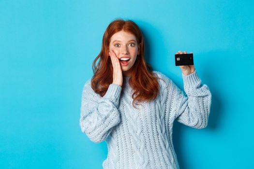 Cute redhead girl in sweater showing credit card, smiling at camera, standing over blue background.