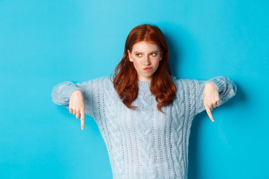 Winter holidays and people concept. Indecisive redhead girl in sweater pointing fingers down and thinking, having doubts, standing over blue background.