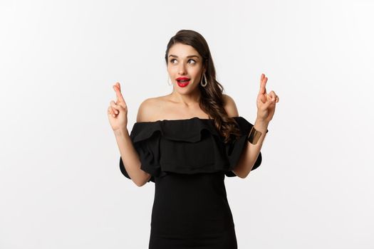 Fashion and beauty. Excited gorgeous woman in black dress, holding fingers crossed and looking at upper left corner, making wish, standing over white background.