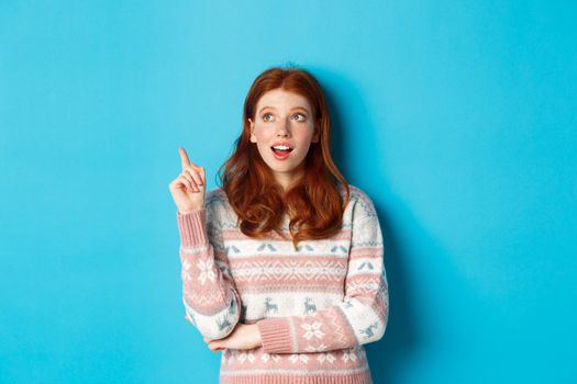 Thoughtful redhead girl in winter sweater having an idea, raising finger in eureka sign and looking up, standing against blue background.