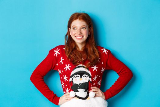 Winter holidays and Christmas Eve concept. Beautiful teenage redhead girl in xmas sweater looking left at logo, smiling pleased, holding hands on waist, blue background.