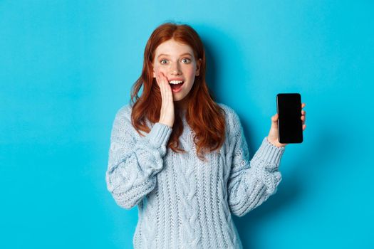 Amazed redhead girl looking at camera, showing smartphone screen, demonstrating mobile application, standing over blue background.