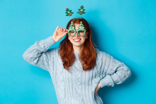 Winter holidays and Christmas sales concept. Beautiful redhead female model celebrating New Year, wearing funny party headband and glasses, smiling at camera.