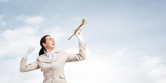 Beautiful woman holding trumpet brass overhead. Young smiling businesslady in white business suit and gloves posing with music instrument on blue sky background. Business concept with musician