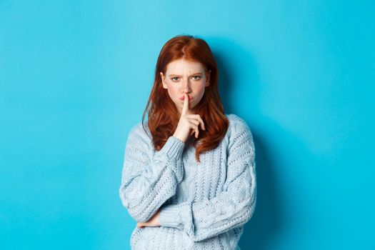 Angry redhead girl hushing at you, show taboo gesture, forbid to speak, standing over blue background in sweater.