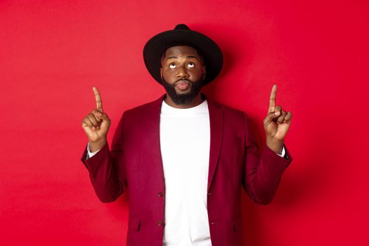 Winter holidays and shopping concept. Curious african american guy in party clothing looking and pointing fingers up, reading promo sign, standing over red background.