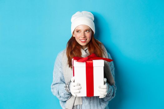 Winter holidays and Christmas sales concept. Thoughtful redhead woman standing in white beanie and gloves, holding holiday gift and looking left, thinking, standing over blue background.