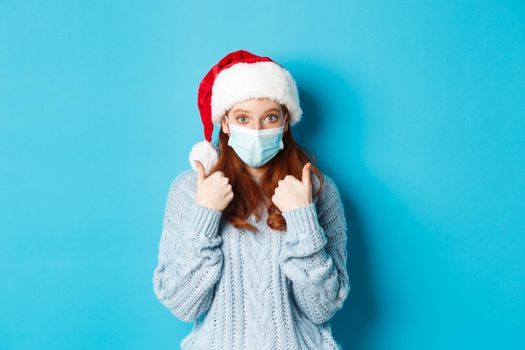 Christmas, quarantine and covid-19 concept. Cute teen redhead girl in santa hat and sweater, wearing face mask from coronavirus, showing thumbs up, standing over blue background.