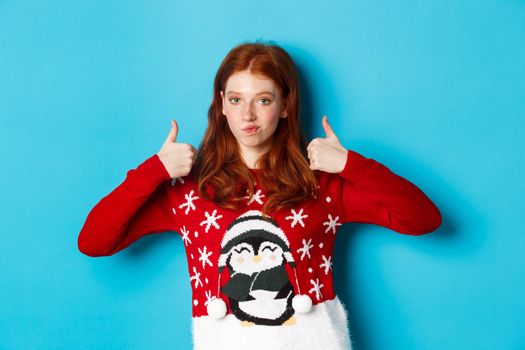 Winter holidays and Christmas Eve concept. Impressed redhead girl in xmas sweater, nod in approval and showing thumb up, praise good product, standing over blue background.