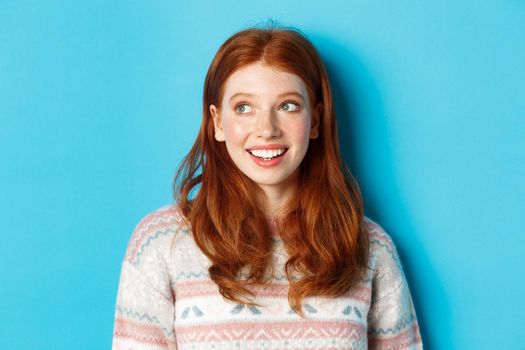 Close-up of dreamy redhead girl in winter sweater looking left, smiling and staring at promo offer, standing against blue background.