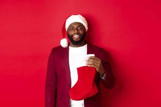 Handsome african american man celebrating winter holidays, holding Christmas sock and smiling, wearing santa hat, standing over red background.
