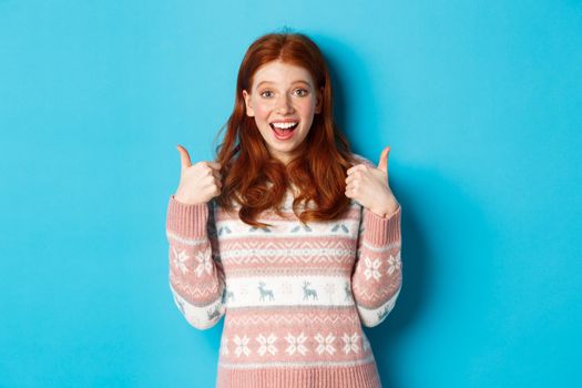 Girl approve something awesome, showing thumb-up, well done gesture and smiling amazed, standing over blue background.