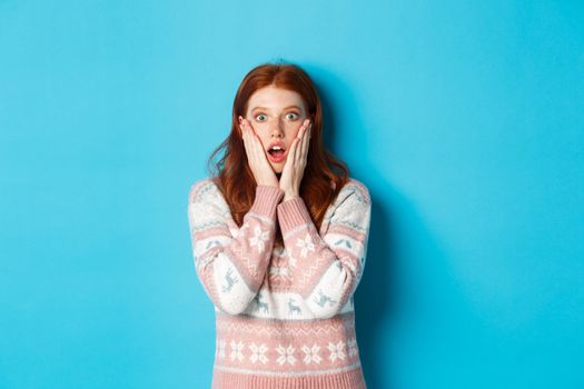 Shocked and surprised redhead girl staring at camera in awe, express complete disbelief, standing against blue background.