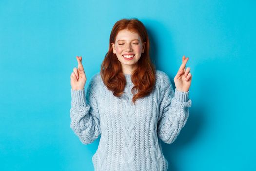 Hopeful redhead girl making a wish, cross fingers for good luck, smiling and anticipating good news or positive result, standing against blue background.