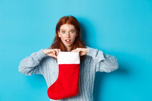 Winter holidays and gifts concept. Funny redhead girl looking surprised, open Christmas stocking and smiling, receiving xmas present, standing against blue background.