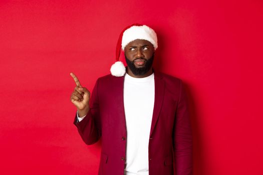 Christmas, party and holidays concept. Skeptical and gloomy african american man pointing finger left, looking disappointed, standing in santa hat against red background.