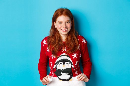 Winter holidays and Christmas Eve concept. Cute redhead girl wearing adorable sweater and smiling, celebrating New Year, standing over blue background.
