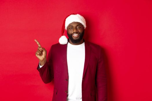 Christmas, party and holidays concept. Handsome Black man in santa hat pointing finger left, showing advertisement, standing happy over red background.