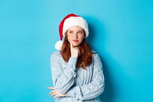 Winter holidays and Christmas Eve concept. Silly redhead girl with freckles, wearing santa hat and thinking, planning New Year celebration, standing over blue background.