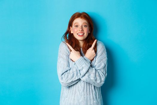 Excited redhead girl pointing fingers sideways, showing two choices and looking tempted at camera, standing against blue background.