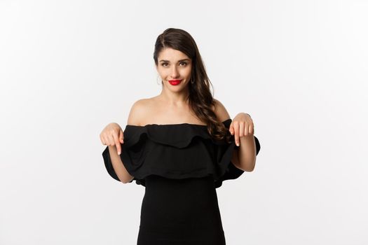 Fashion and beauty. Elegant woman in black dress pointing fingers down, showing promo and smiling, standing over white background.