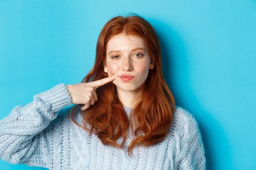 Close-up of sassy teen girl with red hair, pointing at her cheek and staring at camera, standing against blue background.