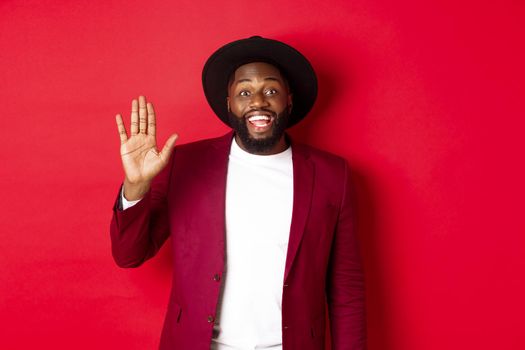 Friendly african american guy waving hand, saying hello and smiling, greeting you, standing over red background.