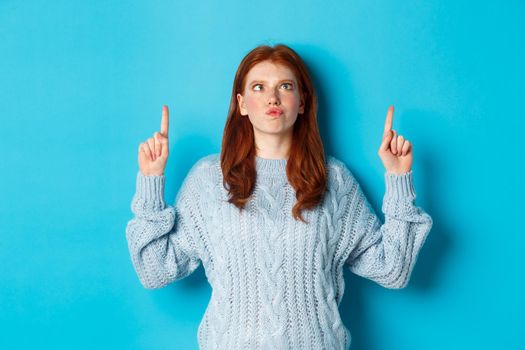 Winter holidays and people concept. Funny and silly redhead girl in sweater, pointing fingers up and squint eyes, standing over blue background.