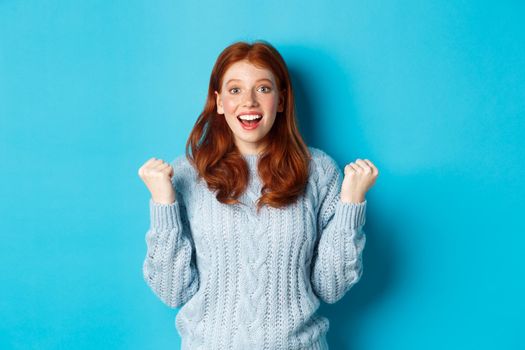 Satisfied redhead girl achieve goal and celebrating, making fist pump gesture and smiling with rejoice, triumphing of win, standing against blue background.
