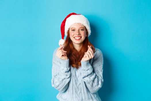 Winter holidays and Christmas Eve concept. Hopeful redhead girl in santa hat, making wish on xmas with fingers crossed, wearing santa hat, standing over blue background.