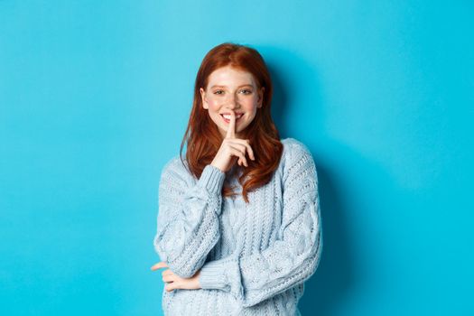 Pretty redhead teenager hushing and smiling, telling a secret, standing in sweater against blue background.