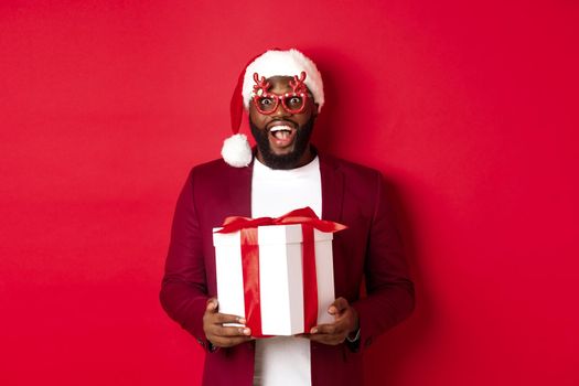 Christmas. Handsome african american man in party glasses and santa hat holding new year gift, looking surprised, receive present and smiling, standing over red background.