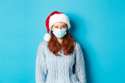Christmas, quarantine and covid-19 concept. Cheeky redhead female model in face mask and santa hat, winking at camera, wishing merry xmas, standing over blue background.