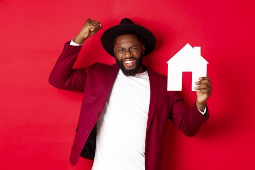 Real estate. Happy Black man celebrating, buying new house, showing paper home model and raising fist up in triumph, standing over red background.