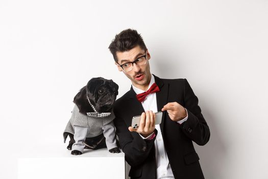 Handsome young man showing something on mobile phone to his dog. Owner shopping online with pet, standing in costumes over white background.