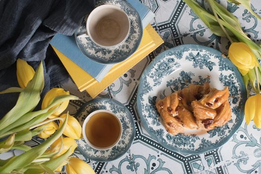 flat lay food, sweet puff, tea in a vintage porcelain cup and a stack of books, a bouquet of yellow withering tulips in a vase,. High quality photo