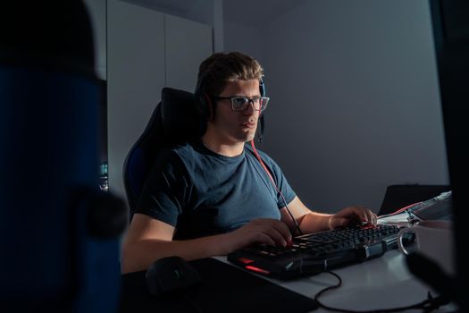 Short blond-haired young man with glasses, cheerful and happy, dressed in black shirt, fond of video games, playing on his laptop computer at night. Young video game player. Dim light from computer monitor, dark room, desk with keyboard, computer screen and coloured lights. Horizontal