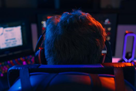 back view of Silhouette of a Young man with short blond hair and glasses, and headphones, wearing a black shirt, playing video games on a computer with headphones. Young gamer illuminated with blue and red coloured lights. Dim light from computer monitor, dark room, desk with keyboard, computer screen and coloured lights. Horizontal.