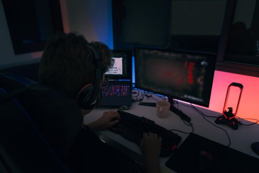 Young man with short blond hair and glasses, cheerful and happy, wearing a black shirt, playing video games on a computer with headphones and using a backlit keyboard. . Dim light from computer monitor, dark room, desk with keyboard, computer screen and coloured lights. Horizontal.