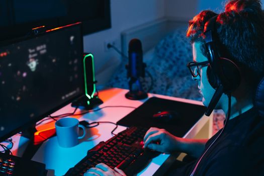 Young man with short blond hair, wearing glasses and headphones, dressed in black shirt, addicted to online video games, playing on a computer with headphones. Young gamer illuminated with blue and red coloured lights. Dim light from computer monitor, dark room, desk with keyboard, computer screen and coloured lights. Horizontal.