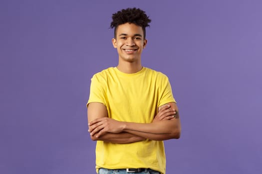 Close-up portrait of confident, smart and professional young male student with dreads, yellow t-shirt, cross arms over chest and smiling pleased, knows what he doing, purple background.