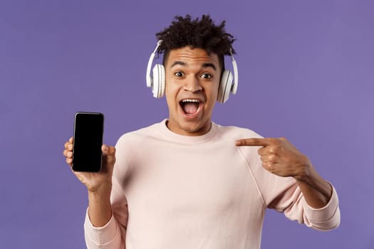 Technology and lifestyle concept. Portrait of excited impressed young man in headphones listening music, online platform or enjoying interesting podcast, pointing at mobile phone, recommending.