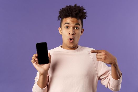 Close-up portrait of excited, cheerful young hispanic male geek talking about his device, new gadget or application, holding mobile phone, pointing at smartphone with amused face.
